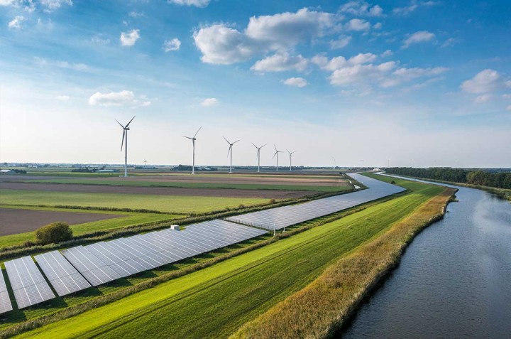 Un extenso campo verde con paneles solares, aerogeneradores, terrenos arados y un río