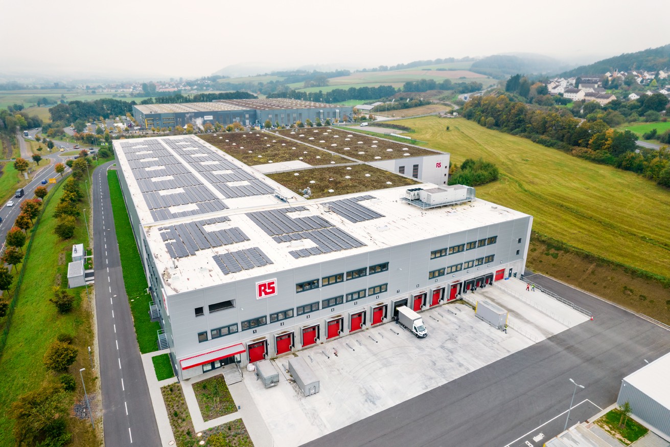 RS Unternehmensbild, das das RS Lager in Bad Hersfeld, Deutschland, aus der Vogelperspektive zeigt. Ein Truck steht vor einer der vielen Laderampen. Im Hintergrund sieht man Felder, Straßen und Landschaft.