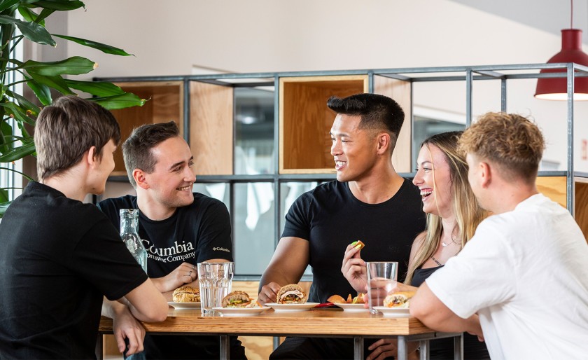 Vier Männer und eine Frau stehen an einem Tisch mit Essen und Getränken und lachen