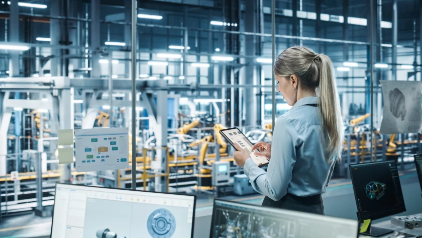 Hands holding a tablet and controlling a robot that's manufacturing a car in the background.