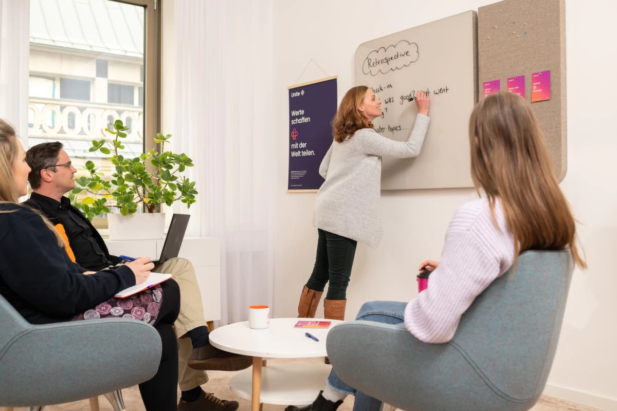 A woman is writing on a whiteboard, three people are sitting on armchairs watching