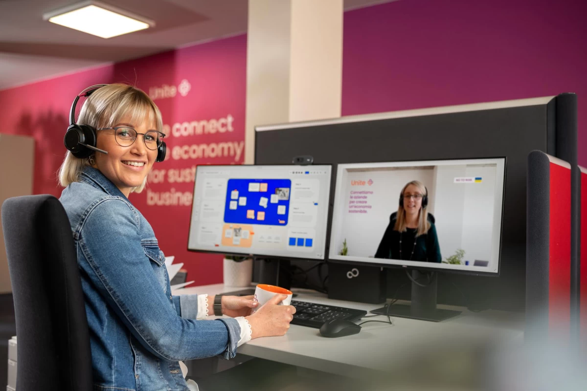 A woman sits in an office in front of a screen and turns to face the camera