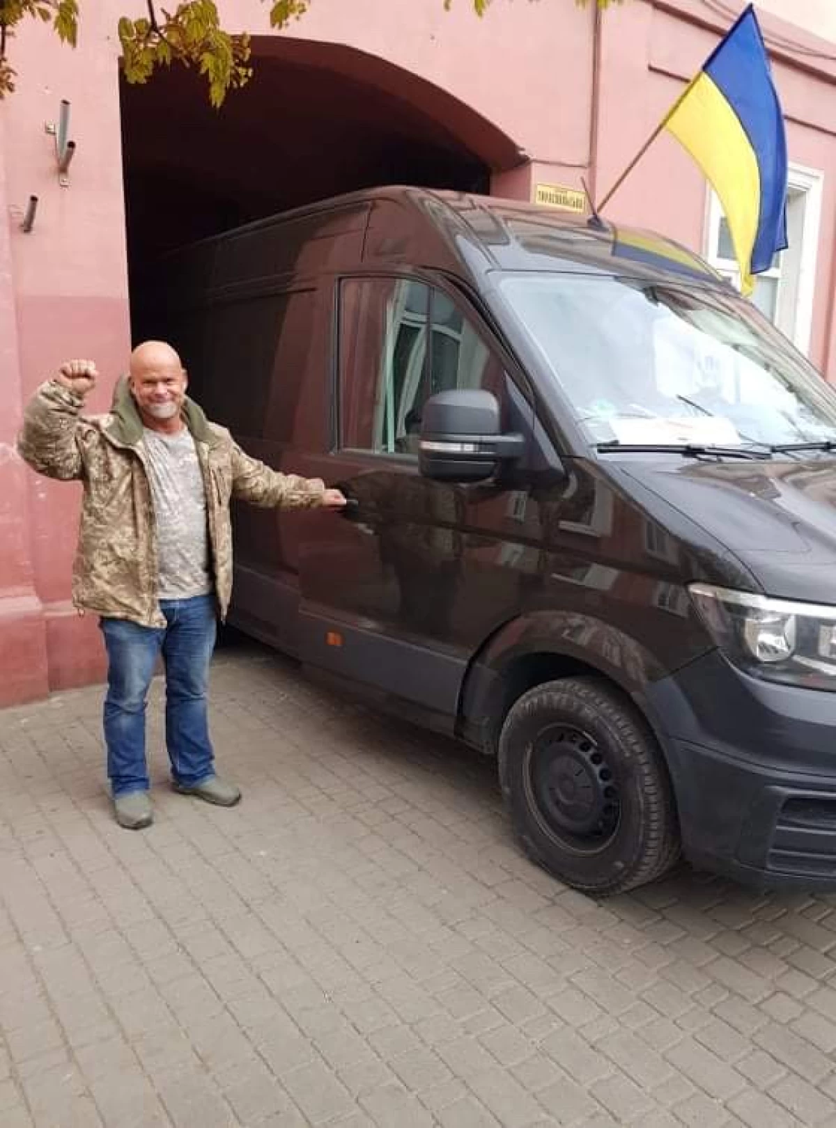 A man stands next to a car and waves his hand in the direction of the photographer