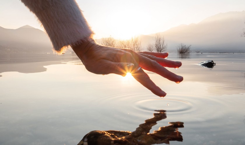 Hand berührt Wasseroberfläche und Sonne reflektiert auf dem Wasser und leuchtet von vorn