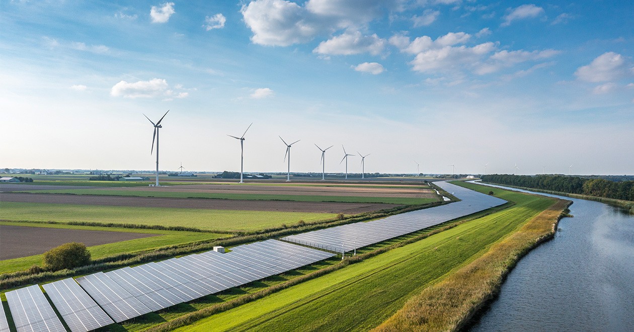 Un extenso campo verde con paneles solares, aerogeneradores, terrenos arados y un río 