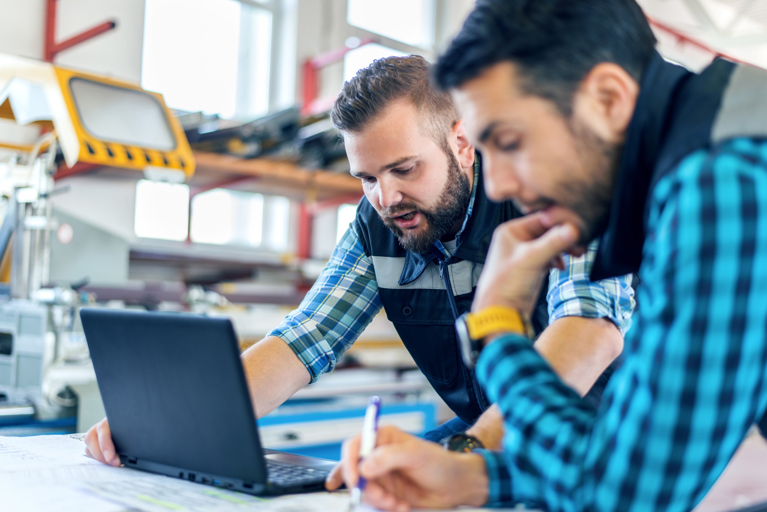 Twee mannen in een fabriek kijken naar een laptop en maken aantekeningen 